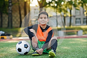 Cute hispanic boy in football uniform with soccer ball after intensive training on stadium at urban park on summer day.