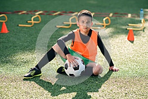 Cute hispanic boy in football uniform with soccer ball after intensive training on stadium at urban park on summer day.