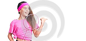 Cute hispanic child girl holding badminton racket pointing thumb up to the side smiling happy with open mouth