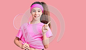 Cute hispanic child girl holding badminton racket smiling happy pointing with hand and finger to the side