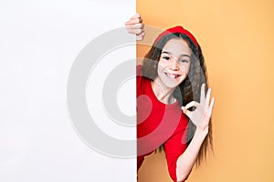 Cute hispanic child girl holding blank empty banner doing ok sign with fingers, smiling friendly gesturing excellent symbol
