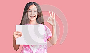 Cute hispanic child girl holding blank empty banner doing ok sign with fingers, smiling friendly gesturing excellent symbol