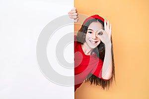 Cute hispanic child girl holding blank empty banner smiling happy doing ok sign with hand on eye looking through fingers