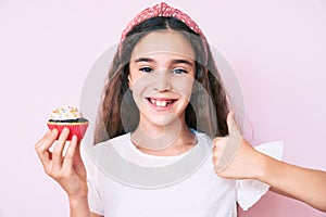 Cute hispanic child girl holding cupcake smiling happy and positive, thumb up doing excellent and approval sign