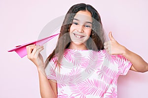 Cute hispanic child girl holding paper airplane smiling happy and positive, thumb up doing excellent and approval sign