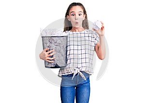 Cute hispanic child girl holding paper bin full of crumpled papers scared and amazed with open mouth for surprise, disbelief face