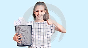 Cute hispanic child girl holding paper bin full of crumpled papers smiling happy and positive, thumb up doing excellent and