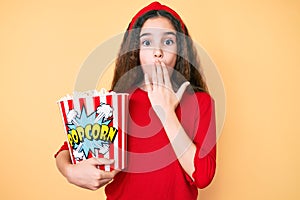 Cute hispanic child girl holding popcorn covering mouth with hand, shocked and afraid for mistake