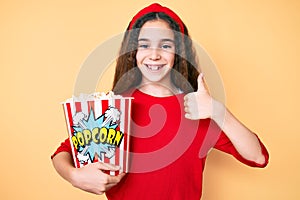 Cute hispanic child girl holding popcorn smiling happy and positive, thumb up doing excellent and approval sign