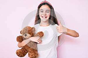 Cute hispanic child girl holding teddy bear smiling happy and positive, thumb up doing excellent and approval sign