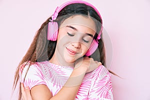 Cute hispanic child girl listening to music using headphones hugging oneself happy and positive, smiling confident