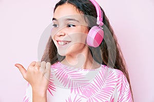 Cute hispanic child girl listening to music using headphones pointing thumb up to the side smiling happy with open mouth