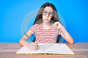 Cute hispanic child girl sitting on the table writing book with angry face, negative sign showing dislike with thumbs down,