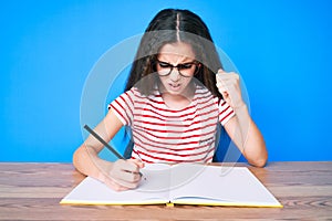 Cute hispanic child girl sitting on the table writing book annoyed and frustrated shouting with anger, yelling crazy with anger