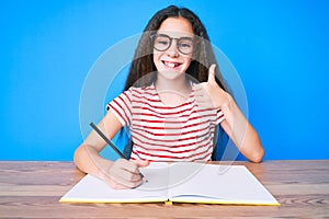 Cute hispanic child girl sitting on the table writing book smiling happy and positive, thumb up doing excellent and approval sign