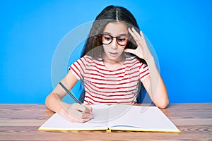 Cute hispanic child girl sitting on the table writing book stressed and frustrated with hand on head, surprised and angry face