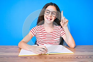 Cute hispanic child girl sitting on the table writing book surprised with an idea or question pointing finger with happy face,