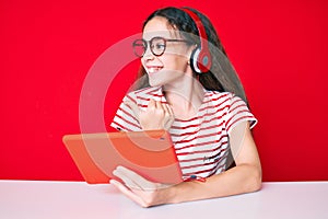 Cute hispanic child girl using touchpad and headphones sitting on the table pointing thumb up to the side smiling happy with open