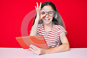 Cute hispanic child girl using touchpad and headphones sitting on the table smiling happy doing ok sign with hand on eye looking