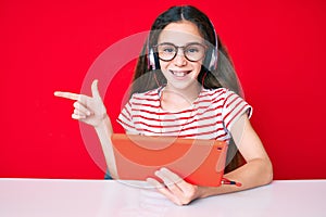 Cute hispanic child girl using touchpad and headphones sitting on the table smiling happy pointing with hand and finger to the