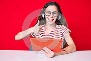 Cute hispanic child girl using touchpad and headphones sitting on the table smiling happy and positive, thumb up doing excellent