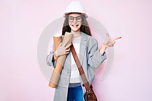 Cute hispanic child girl wearing architect hardhat holding blueprints smiling happy pointing with hand and finger to the side