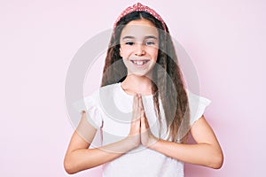 Cute hispanic child girl wearing casual clothes and diadem praying with hands together asking for forgiveness smiling confident