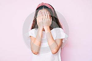 Cute hispanic child girl wearing casual clothes and diadem with sad expression covering face with hands while crying
