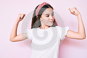 Cute hispanic child girl wearing casual clothes and diadem showing arms muscles smiling proud