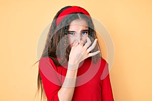 Cute hispanic child girl wearing casual clothes and diadem smelling something stinky and disgusting, intolerable smell, holding