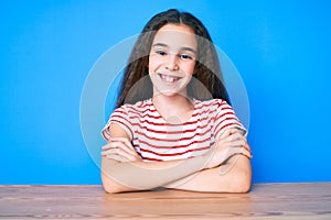 Cute hispanic child girl wearing casual clothes sitting on the table happy face smiling with crossed arms looking at the camera