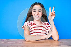 Cute hispanic child girl wearing casual clothes sitting on the table smiling with happy face winking at the camera doing victory