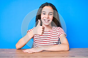 Cute hispanic child girl wearing casual clothes sitting on the table smiling happy and positive, thumb up doing excellent and