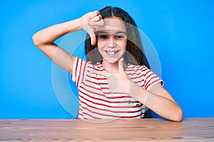 Cute hispanic child girl wearing casual clothes sitting on the table smiling making frame with hands and fingers with happy face