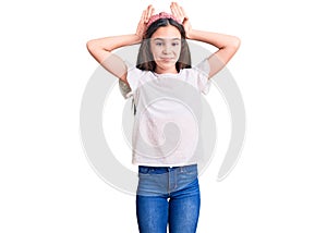 Cute hispanic child girl wearing casual white tshirt doing bunny ears gesture with hands palms looking cynical and skeptical