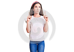 Cute hispanic child girl wearing casual white tshirt pointing up looking sad and upset, indicating direction with fingers, unhappy
