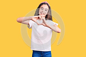 Cute hispanic child girl wearing casual white tshirt smiling in love doing heart symbol shape with hands