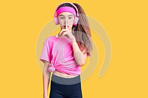 Cute hispanic child girl wearing gym clothes and using headphones asking to be quiet with finger on lips