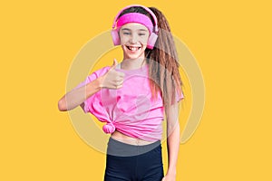 Cute hispanic child girl wearing gym clothes and using headphones doing happy thumbs up gesture with hand