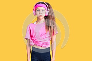 Cute hispanic child girl wearing gym clothes and using headphones puffing cheeks with funny face