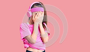 Cute hispanic child girl wearing gym clothes and using headphones with sad expression covering face with hands while crying