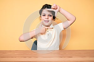 Cute hispanic child wearing casual clothes sitting on the table smiling making frame with hands and fingers with happy face