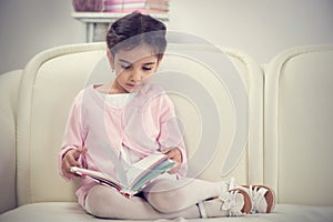 Cute hispanic little girl reading book on couch