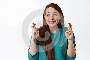 Cute hopeful redhead woman cross fingers for good luck, smiling and anticipating news, looking aside and waiting for