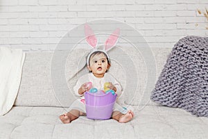 Cute Indian baby girl with pink bunny ears and basket of colorful eggs celebrating Easter holiday