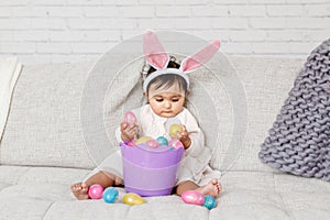 Cute Indian baby girl with pink bunny ears and basket of colorful eggs celebrating Easter holiday