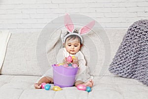 Cute Indian baby girl with pink bunny ears and basket of colorful eggs celebrating Easter holiday