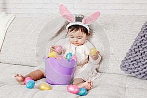 Cute Indian baby girl with pink bunny ears and basket of colorful eggs celebrating Easter holiday