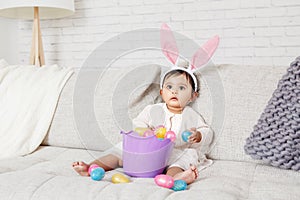 Cute Indian baby girl with pink bunny ears and basket of colorful eggs celebrating Easter holiday