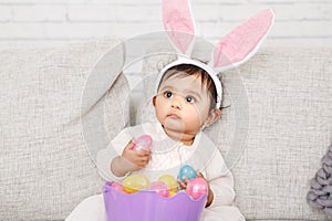 Cute Indian baby girl with pink bunny ears and basket of colorful eggs celebrating Easter holiday